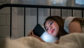 Asian young beautiful woman using mobile phone at night in bedroom. Attractive girl lying down on bed, feeling happy and relax while watching movie and swiping chatting on smartphone at home.