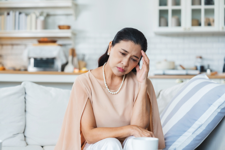 Upset Asian senior woman sitting on sofa, lost in thoughts, feeling lonely, disappointed, alone at home, unhappy, anxious, missing or sad. Senior woman experiencing mental health problems