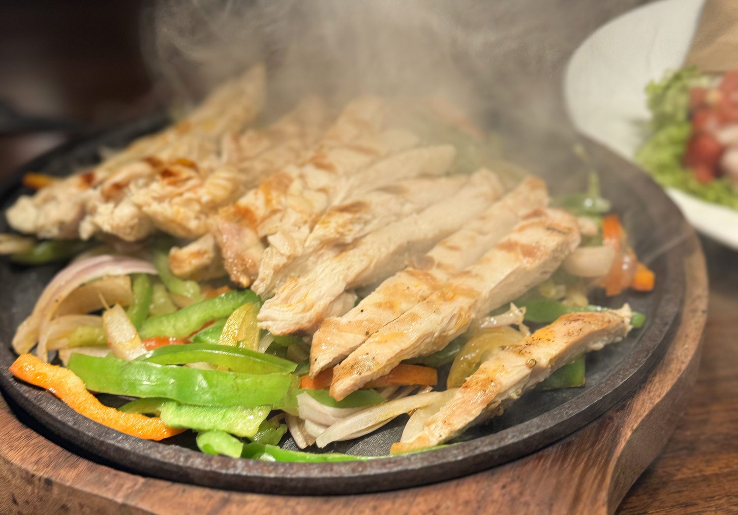 Image of sizzling chicken fajitas in an iron skillet/ with sauteed peppers and onions, steaming hot on a wooden board, enhanced with chilies