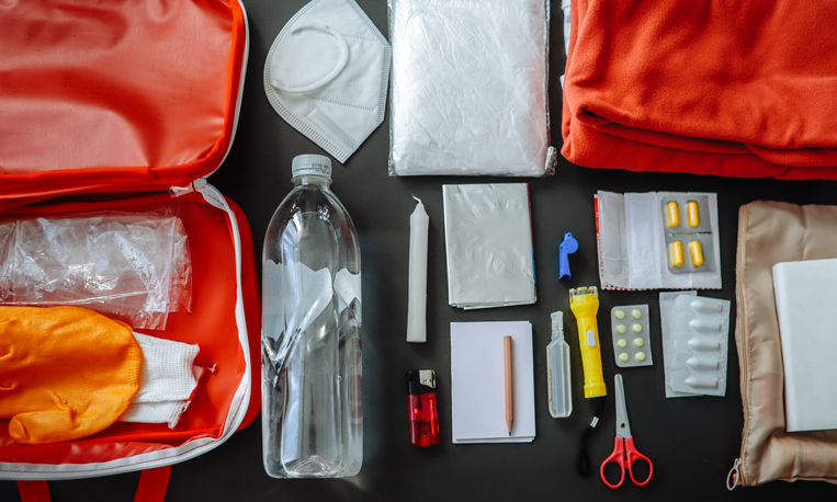 Doomsday prepping: water bottle, protective mask, candle, lighter, warm clothing, scissors, packing bags, pills, flashlight. Emergency kit, top view in a real home setting. Banner top view