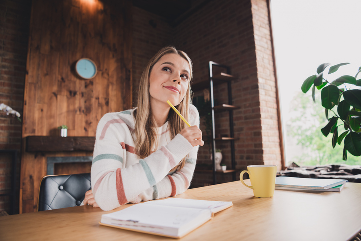 Pensive young woman in cozy sweater daydreams while writing at home, surrounded by natural light in her charming living room
