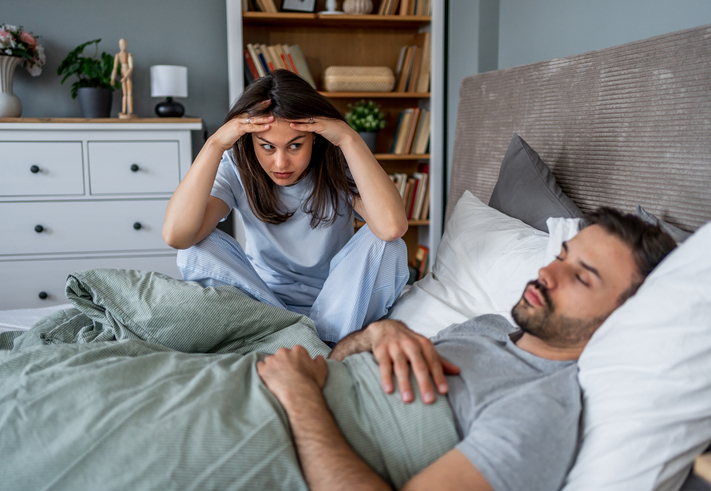 Frustrated woman sitting on bed while man sleeping peacefully