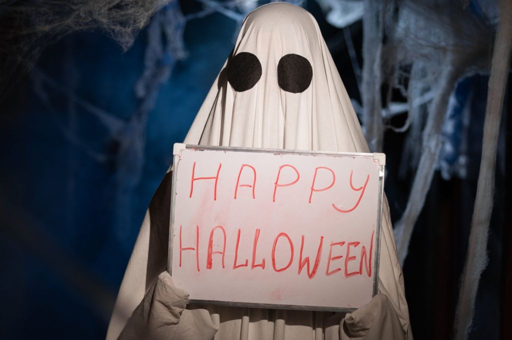 Funny ghost in dark corridor with cobweb showing board with happy halloween inscription positive expression. Happy halloween spooky