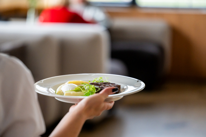 Close-up of a hand holding a plate with poached egg, meat, mashed potatoes, and greens in a modern restaurant setting. Blurred background with people and furniture