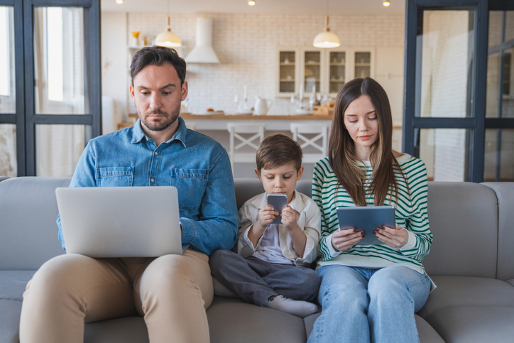Caucasian family watching gadgets while sitting on sofa at home. Dad, mom and son using digital devices tablet laptop phone, everyone using their own gadget. Social networks dependence concept