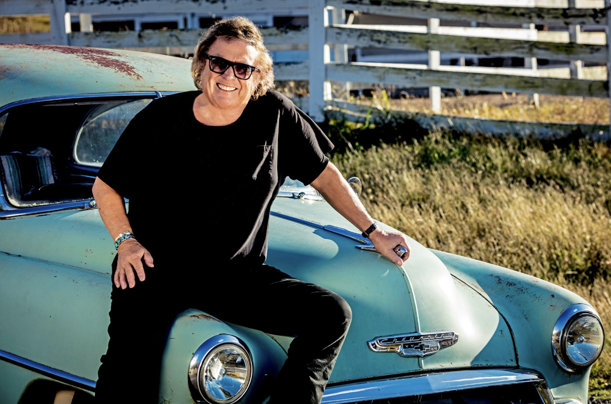A smiling man in sunglasses sitting on the hood of a vintage turquoise car in a grassy outdoor setting.